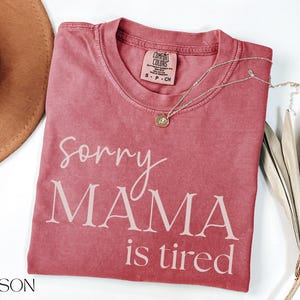 May include: Crimson-colored t-shirt with the text "Sorry MAMA is tired" in white script. A gold necklace with a pendant rests on the shirt. A brown hat and dried leaves are also in the image.