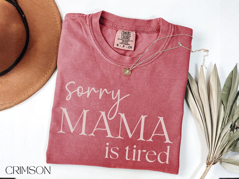 May include: Crimson-colored t-shirt with the text "Sorry MAMA is tired" in white script. A gold necklace with a pendant rests on the shirt. A brown hat and dried leaves are also in the image.