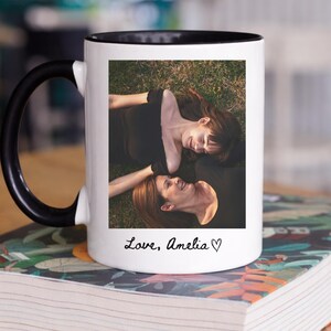 May include: White ceramic mug with a black handle and rim, featuring a photograph of two people lying on grass. The mug has the text "Love, Amelia" in a handwritten font, with a heart symbol.