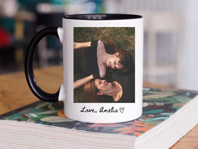 May include: White ceramic mug with a black handle and rim, featuring a photograph of two people lying on grass. The mug has the text "Love, Amelia" in a handwritten font, with a heart symbol.