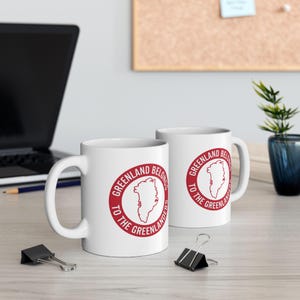 Könnte beinhalten: Zwei weiße Keramikbecher mit einem roten kreisförmigen Design mit der Aufschrift "GREENLAND BELONGS TO THE GREENLANDERS" um eine Karte von Grönland. Die Becher stehen auf einer hellen Oberfläche mit einem Laptop und Bürobedarf im Hintergrund.