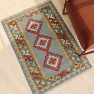 May include: A rectangular area rug featuring a light blue center field with three prominent red and white diamond-shaped geometric motifs. The border displays a repeating pattern of green, red, and yellow shapes with short fringe on the ends.