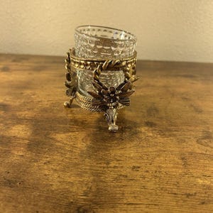 Puede incluir: Un portavelas de estilo vintage con una copa de cristal transparente y adornos de metal dorado. La base metálica presenta diseños florales y de hojas, añadiendo un toque de elegancia. El portavelas está diseñado para una pequeña vela votiva.