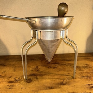 May include: A silver-coloured metal food mill with a conical perforated strainer, a wide circular rim, and a long handle. The device is supported by three curved metal legs and includes a dark brown wooden pestle resting inside the basin.
