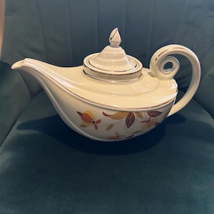 May include: A white ceramic teapot with a curved spout and handle. The teapot features a lid with a finial and is decorated with a floral pattern in shades of orange, yellow, and brown. The rim and handle have a gold trim.