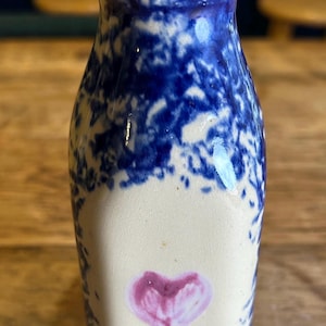 May include: A small, ceramic vase with a white base and blue splatter pattern. A pink heart is painted on the front. The vase has a narrow neck and a slightly flared rim. Decorative pottery.