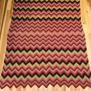 Può includere: Una coperta rettangolare all'uncinetto con un motivo a chevron ripetitivo. Il design è composto da strisce orizzontali in varie tonalità di rosa, dal rosa chiaro al magenta intenso, accentuate da sottili linee di verde scuro e nero.