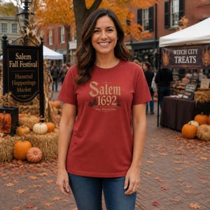 Könnte beinhalten: Ein ziegelrotes Kurzarm-T-Shirt mit dem goldenen Schriftzug Salem 1692 und dem Satz They Missed One sowie einer kleinen Grafik eines Hauses und Bäumen, die mittig auf der Brust aufgedruckt sind.