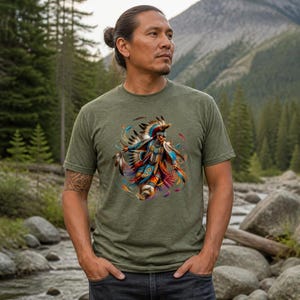 May include: An olive green short-sleeved t-shirt featuring a colorful graphic print of a person in traditional regalia with a feathered headdress. The design includes vibrant blue, orange, and yellow accents, showcasing intricate patterns and feathers.