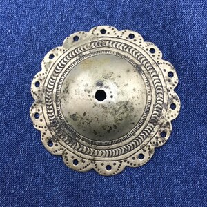 May include: A circular, tarnished brass-toned metal disc featuring a raised, domed center with a small hole. The outer edge has a scalloped design with small punched holes, and the surface is decorated with engraved, repeating linear patterns.