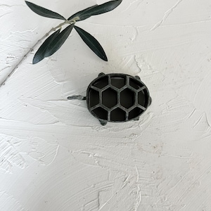 May include: A dark grey metal turtle-shaped container with a honeycomb design on its back. The container has small legs and a head. A sprig of green leaves is in the background.