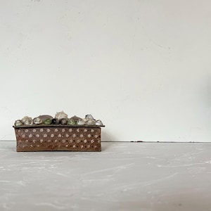 May include: A rectangular, brown metal box with a textured surface and a lid decorated with clear and green glass stones. The box is set against a neutral background, highlighting its rustic design.