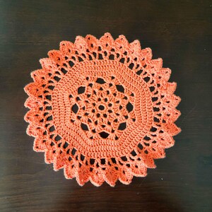 May include: A round, bright orange crochet doily featuring a central star-like pattern surrounded by concentric rows of intricate stitching and a decorative, scalloped edge with pointed, petal-shaped loops forming the outer border.