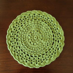 May include: A round, handmade crochet doily in a light green color. The doily features a detailed, openwork pattern with a scalloped edge. The circular design is centered and radiates outwards, creating a decorative and textured piece.