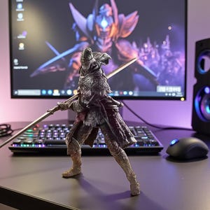 Puede incluir: Figura detallada de un guerrero con armadura ornamentada, sosteniendo una espada larga. La figura está frente a un monitor de computadora que muestra un personaje de fantasía. También se ven un teclado, un ratón y un altavoz.