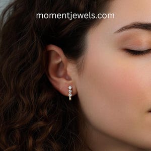 May include: A close-up of a woman's ear wearing a gold earring with three clear, round gemstones. The earring is a small hoop design. The website momentjewels.com is visible in the image.