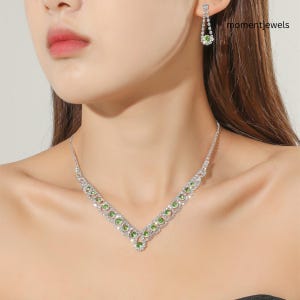 Op de afbeelding: Een set bestaande uit een V-vormige ketting en bijpassende oorbellen. De sieraden zijn versierd met heldere, rondgeslepen kristallen en kleine, levendig groene ronde stenen die in een zilverkleurig metalen frame zijn gezet voor een geheel.