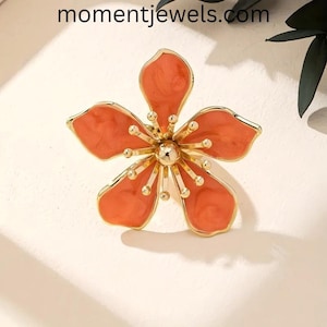 May include: A gold-toned flower-shaped brooch with five petals in a coral hue. The brooch has a central gold sphere and smaller gold accents. The website momentjewels.com is visible at the top of the image.