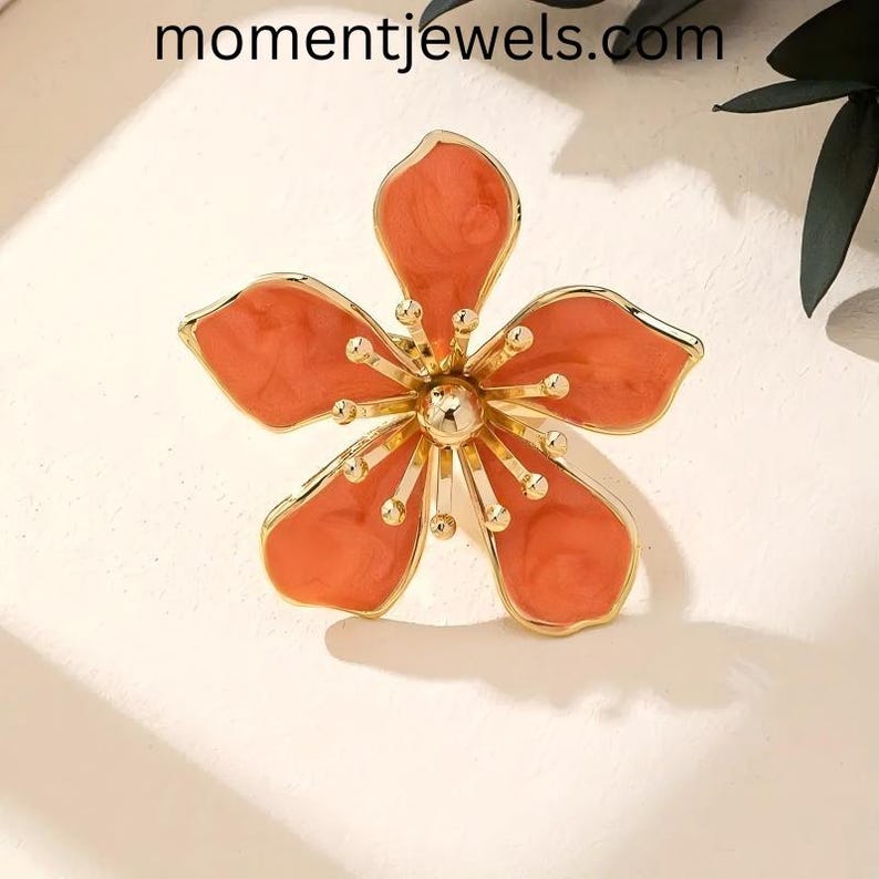May include: A gold-toned flower-shaped brooch with five petals in a coral hue. The brooch has a central gold sphere and smaller gold accents. The website momentjewels.com is visible at the top of the image.