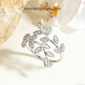 Puede incluir: Un anillo plateado con diseño de hojas, adornado con pequeñas gemas brillantes. El anillo se envuelve alrededor del dedo, con múltiples elementos en forma de hoja. El sitio web momentjewels.com es visible.