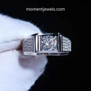 May include: A silver ring featuring a large, clear, square-cut gemstone at its centre. The band is embellished with numerous smaller, sparkling stones. The website momentjewels.com is visible. This ring is a piece of jewellery.
