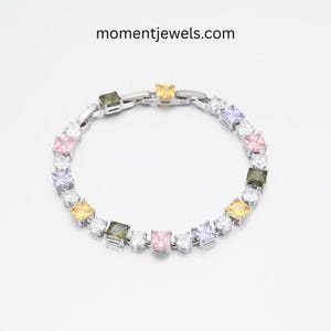Op de afbeelding: Een zilveren armband met vierkante, veelkleurige edelstenen, waaronder roze, geel, groen en paars, afgewisseld met heldere, ronde stenen. De armband wordt getoond op een witte achtergrond, met de tekst "momentjewels.com" erboven.