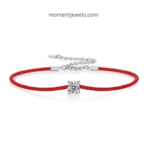 May include: A red cord bracelet with a silver clasp and a single, round, clear gemstone. The bracelet is adjustable and features the text "momentjewels.com".