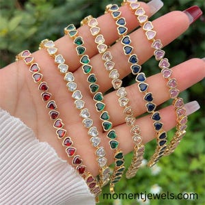 Pode incluir: Uma mão exibe várias pulseiras douradas, cada uma adornada com pedras preciosas em forma de coração em várias cores, incluindo vermelho, transparente, verde, azul e rosa. As pulseiras estão dispostas em uma fileira, mostrando os diferentes tons das pedras preciosas.