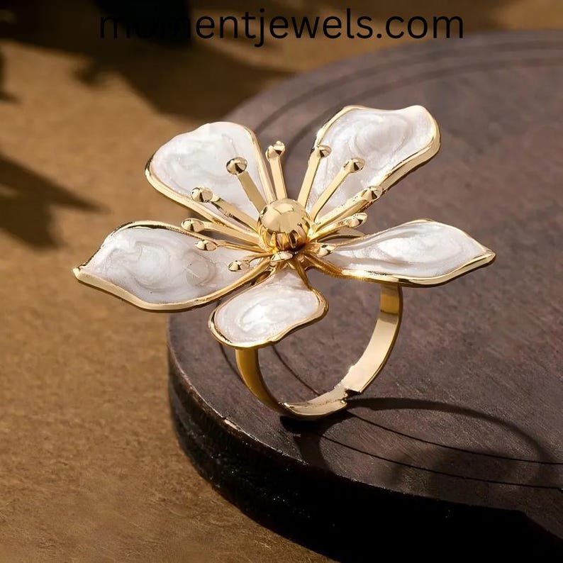 May include: A gold-toned ring featuring a large, stylized flower design. The flower has five white petals with a marbled effect and a gold center. The ring is set on a dark wooden surface.