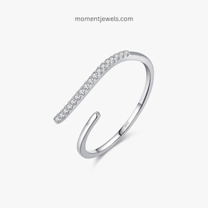 May include: A silver-colored ring with a curved design. The ring features a row of small, clear stones set along a straight bar. The website momentjewels.com is printed on the top of the image.