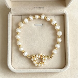 Può includere: Un bracciale con perle d'acqua dolce bianche e piccole perline color oro disposte in un motivo alternato. Il gioiello presenta una chiusura a T con un anello circolare decorato con ulteriori piccole perle. Il testo momentjewels è visibile.