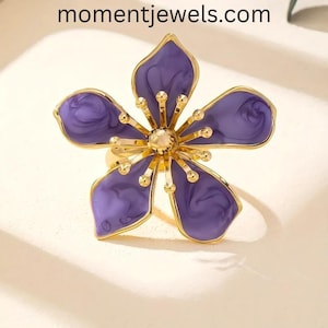May include: A gold-toned flower ring with five purple petals and a gold center. The petals have a marbled effect. The ring is a floral design. The website momentjewels.com is visible in the image.