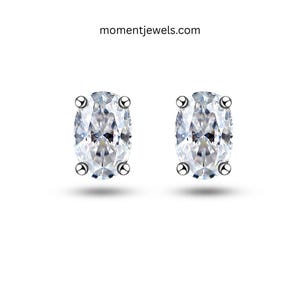 May include: A pair of oval-cut clear stud earrings with four-prong settings in a metallic finish. The text momentjewels.com is visible at the top centre. These earrings feature faceted stones that reflect light, creating a bright, clear appearance.