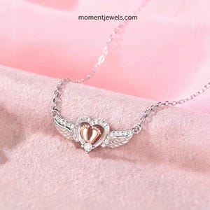 May include: Silver necklace with a heart-shaped pendant featuring baby footprints and angel wings. The pendant is embellished with small, clear crystals and a single, larger crystal. The website momentjewels.com is visible in the image.