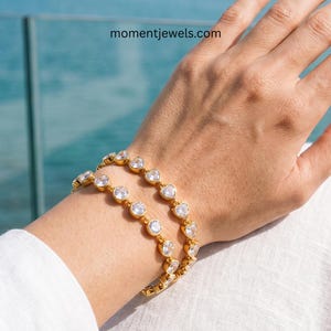 May include: Two gold bracelets with clear, round and heart-shaped gemstones. The bracelets are worn on a wrist, with the website momentjewels.com visible in the background.
