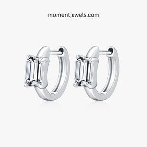 May include: A pair of silver hoop earrings, each featuring a rectangular, clear gemstone. The earrings are set against a white backdrop. The website momentjewels.com is visible at the top of the image.