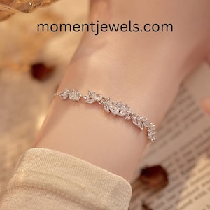 Könnte beinhalten: Ein filigranes silbernes Armband mit floralem Design und klaren Edelsteinen. Das Armband wird am Handgelenk getragen, wobei die Website momentjewels.com sichtbar ist. Ein schönes Schmuckstück.