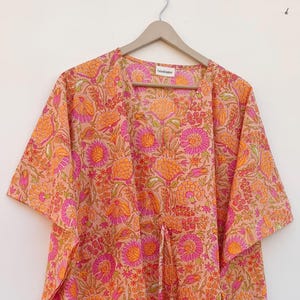 May include: A long, flowing kaftan with a floral pattern. The fabric is a light peach colour with a repeating design of orange and pink flowers and green leaves. The kaftan has short sleeves and a drawstring waist.