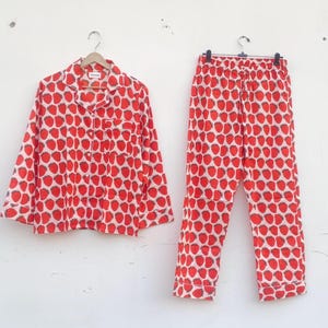Puede incluir: Un conjunto de pijama de dos piezas con un estampado de fresas rojas sobre fondo blanco. El conjunto incluye una camisa con botones y un pantalón a juego. La camisa tiene cuello y bolsillo en el pecho. El pantalón tiene cintura elástica.