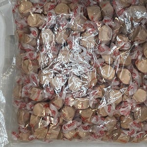 May include: A clear plastic bag filled with individually wrapped, tan-colored taffy candies. The wrappers have a red and white design. The candies are tightly packed together, creating a full, textured appearance.