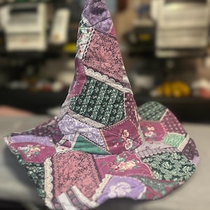 May include: A patchwork witch hat with a wide brim. The hat is made of various fabrics in shades of purple, green, and pink, with floral and lace patterns. The hat has a pointed top and a quilted texture.