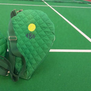 May include: Green quilted tennis racket bag with a yellow tennis ball graphic and monogrammed initials. The bag has a shoulder strap and gold-colored hardware. The bag is on a green surface with white lines.