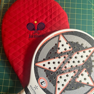May include: A red quilted paddle with the name "Jansen" and a tennis racket embroidered on it, next to a white and black paddle with a red star design and the text "Master Athletics" and "Edge".