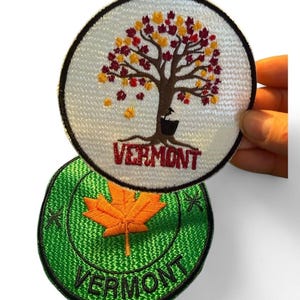May include: Two round embroidered patches. The top patch features a tree with red and yellow leaves, the word "VERMONT" in red, and a black border. The bottom patch is green with an orange maple leaf and the word "VERMONT".