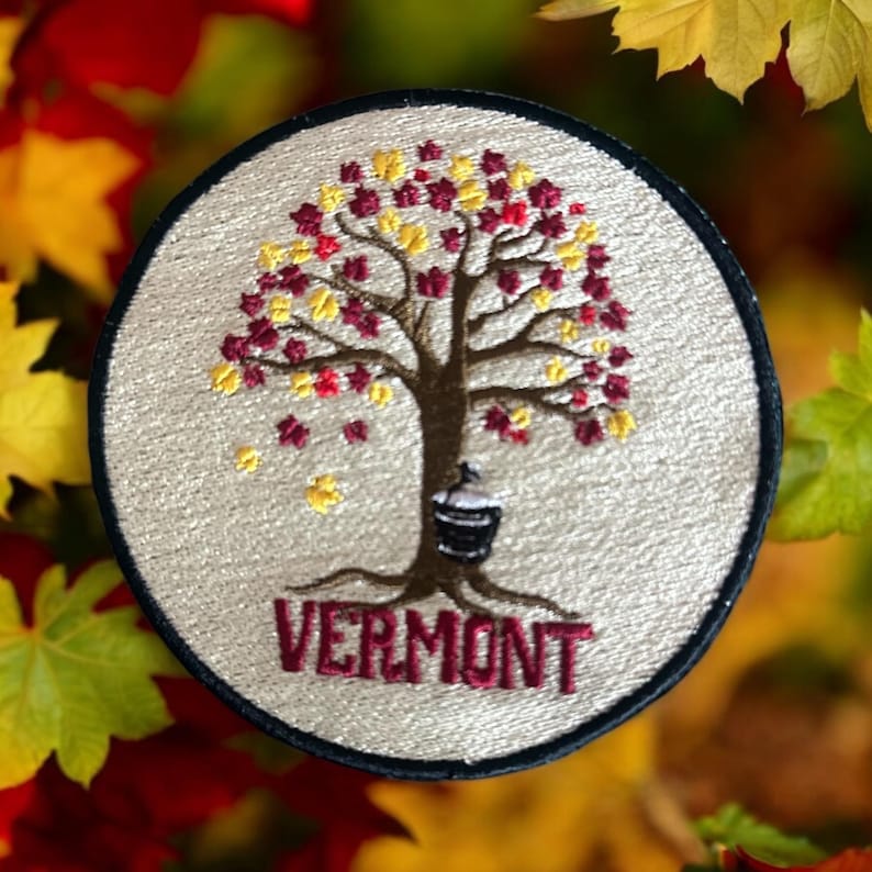 Pode incluir: Patch bordado circular com uma &aacute;rvore com folhas vermelhas e amarelas, um balde e a palavra "VERMONT" em bord&ocirc;. O patch tem um fundo creme e uma borda preta, em um fundo de folhagem de outono.