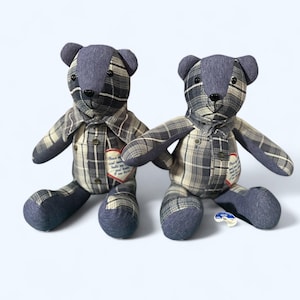 May include: Two handmade teddy bears with plaid shirts and denim accents. The bears have button closures and small heart-shaped patches with text. The bears are sitting on a white surface.