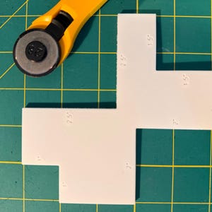 May include: A white, L-shaped template with measurements in inches, next to a yellow rotary cutter. The template has dimensions of 2.5" x 2.5" and 1" x 2". The background is a green cutting mat with a grid pattern.