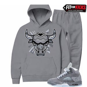 May include: A gray hoodie and sweatpants set with a bull graphic on the front. The hoodie has a drawstring hood and a kangaroo pocket. Gray and white sneakers with the number 23 are also in the image. The text "FIT FOR KICKS" is in the top right corner.