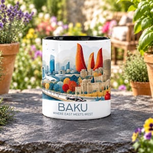 Op de afbeelding: Een witte keramische mok met een zwarte binnenkant voorzien van een kleurrijke illustratie van de skyline van Bakoe, inclusief de Flame Towers, historische stenen muren en een racewagen. De tekst BAKU WHERE EAST MEETS WEST staat in het blauw op de voorzijde.