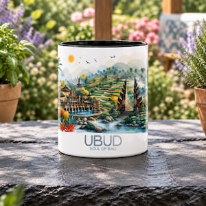 Könnte beinhalten: Ein weißer Keramikbecher mit schwarzem Innenrand, der eine farbenfrohe Illustration einer balinesischen Landschaft mit Reisterrassen, Tempeln und Wasser zeigt. Der Text UBUD SOUL OF BALI ist in Grau am unteren Rand des Bechers aufgedruckt.